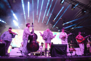 Band performs on stage with multiple musicians and bright blue spotlights overhead; central singer wearing a fluffy outfit and others playing guitar, violin, and drums.