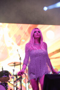 Female singer on a brightly lit stage wearing a glittery purple dress, with drums and colorful backdrop behind her.