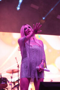 Female singer on stage under pink lighting, reaching out with microphone in hand.]?