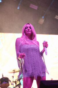 Female singer on stage wearing a glittery pink dress, holding a white microphone, bathed in purple lighting with a drum set in the background.