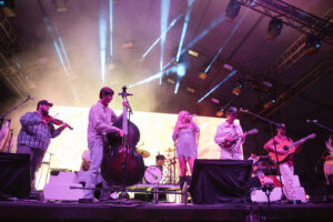 Live band on stage performing with violinist, bassist, singer, guitarist, and drummer under bright stage lights.