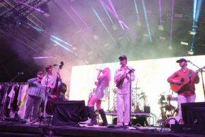 A live band performs on a lit stage with purple and blue spotlights, featuring a singer center stage, guitarists and a bassist, and a drummer in the background.