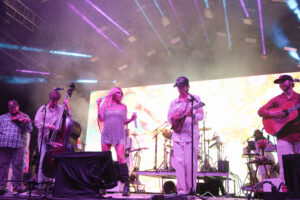 Band on stage performing live: female singer at center with microphone, guitarists and a violinist flanking her, colorful stage lights and smoky atmosphere behind.