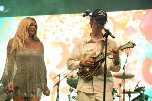 Female singer in a silver sequined dress stands at a microphone beside a man playing a guitar on a brightly lit stage with colorful background.”