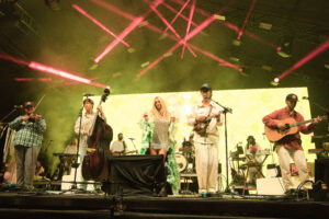 Live band on stage performing with guitars, violin, and drums under pink laser lights.