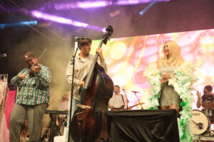 Live band on stage with a bassist center, violinist left, and a singer in a feather boa on the right, colorful lights in the background.