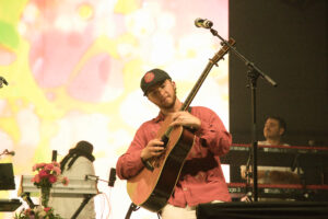 Guitarist in a pink shirt and cap playing an acoustic guitar on a brightly lit stage with a colorful backdrop.