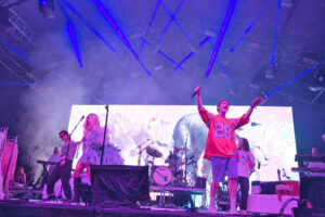Live concert on a large stage with purple lighting; a vocalist in a red jersey raises a microphone amid band members and bright screens behind.