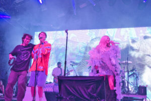 Live concert with three performers on stage under purple-blue lights with smoke and a bright LED screen behind them.