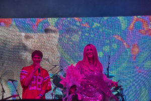Two performers sing on a concert stage in front of a bright, colorful LED backdrop; a male singer on the left and a woman in a pink sequined dress on the right, with drums behind them.