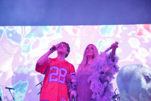 Two singers perform on stage holding microphones, with a colorful abstract backdrop behind them.