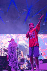 Male singer in a red 88 jersey sings into a microphone on a lit stage, arm raised toward the crowd; drummer and a fluffy feather prop are in the background.