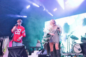 Male singer in a red sports jersey performs on stage beside a female singer in a sparkly dress and feather boa under blue stage lights.