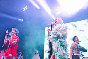 Two singers perform on a brightly lit stage; woman center sings into a microphone wearing a white feathered outfit