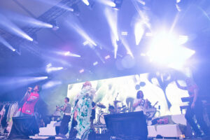 Concert stage with multiple performers under bright blue and white lights, a large screen displaying abstract visuals in the background.