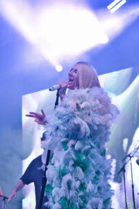 Female singer on stage, holding a microphone and wearing a white feathered costume under bright stage lights.