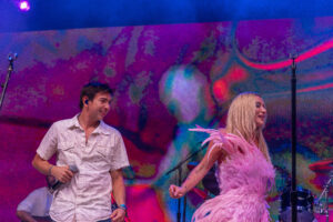 Two performers on a colorful stage: a man holding a microphone in a light patterned shirt, and a woman in a pink feathered outfit dancing before a vibrant LED backdrop.