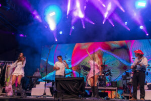 Live band on stage under blue and purple lights with colorful LED backdrop and performers playing instruments, frontwoman in white dress on left.