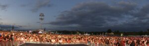 Large outdoor concert crowd at dusk, facing a distant stage with lights and a cloudy sky above.
