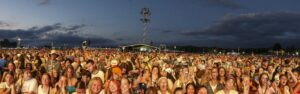 Massive outdoor music festival crowd at dusk, with a distant stage and tall lighting rig above the audience.