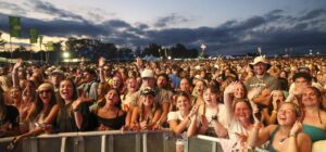 Large crowd at an outdoor concert, people at the barrier waving and cheering as the sun sets behind them.