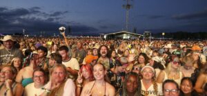 Large crowd of festival goers at dusk with a distant stage and lighting rigs visible in the background.
