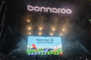 Stage at Bonnaroo festival with a giant LED screen displaying colorful graphics and the festival name above a bustling concert crowd.
