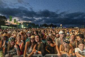 Large crowd of festivalgoers at dusk, standing behind a barrier with flags and lights in the background, smiling and looking toward the stage.
