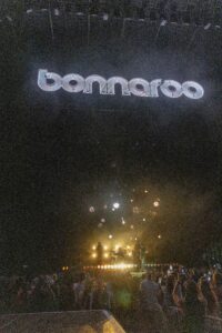 Night-time Bonnaroo concert: a band on stage with bright lights and smoke, fireworks blooming over a cheering crowd.",