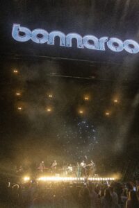 Night concert at Bonnaroo with a large illuminated 'bonnaroo' sign above the stage; crowd holds up phones.