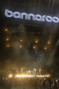 Concert crowd watching a live band on a well-lit stage with a large illuminated Bonnaroo sign overhead.