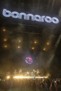 Nighttime concert with a large 'bonnaroo' sign overhead and a band performing on stage, crowd cheering below.