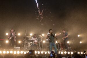 Band performs on a dimly lit stage with guitars and drums; bright round stage lights and a crowd in the foreground, fireworks in the night sky above.