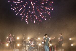 Band performs on a lit stage with guitars and drums as bright fireworks explode overhead at nightfall.