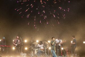 Band performs on stage at night with bright stage lights and a pink fireworks display above them.