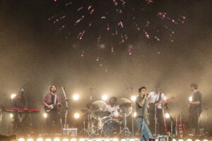A live band performs on a smoky stage with guitars, drums, and a keyboard under bright lights and a rising crowd.