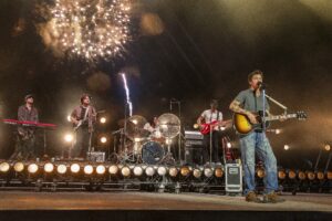 Live concert scene: lead singer with a sunburst guitar at the front of a stage, other band members on keyboards, guitars and drums, with fireworks in the night sky behind them.",