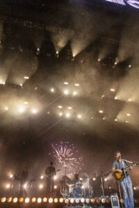Male singer with guitar performing on a smoky concert stage, bright orange lights and a fireworks graphic in the background.
