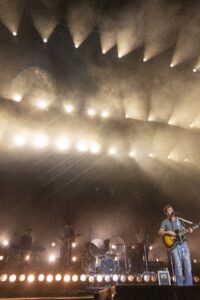 Band performs on a foggy, illuminated stage; guitarist sings into a mic in the foreground as other musicians play in back.