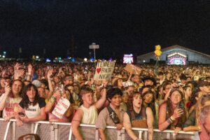 Crowd at a nighttime outdoor concert behind a metal barrier, many people waving hands and holding signs and phones near a lit stage in the distance.