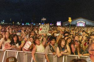 Large nighttime concert crowd behind a barrier, many people holding signs and upraised phones toward a distant stage.
