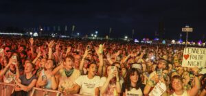 Dense nighttime concert crowd with raised hands and illuminated phone screens, festival tents and colorful lights in the background.