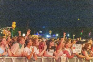 Crowd at a nighttime concert, hands in the air behind a barrier with signs visible in the foreground.