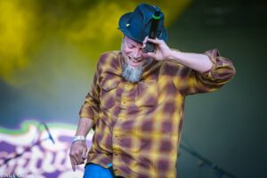 Man with a gray beard wearing a yellow plaid shirt and a blue hat sings into a handheld microphone on stage.
