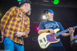 Two male musicians performing on a stage: one wearing a yellow plaid shirt holding a microphone, the other in a blue cap playing a cream-colored bass guitar with sunglasses and ear monitors.