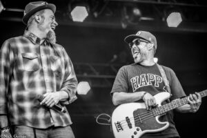 Two male musicians on stage: a bearded man in a plaid shirt and hat laughing, and a cap-wearing, sunglasses-clad bass guitarist playing a white bass.