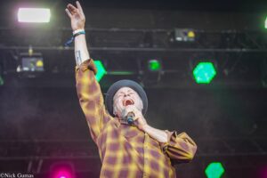 Male singer on stage wearing a plaid shirt and hat, raising one arm while singing into a microphone with colorful stage lights behind him.