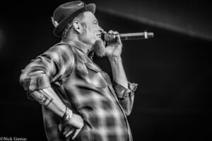 Male performer wearing a hat and plaid shirt, singing into a handheld microphone on stage (black and white photo).