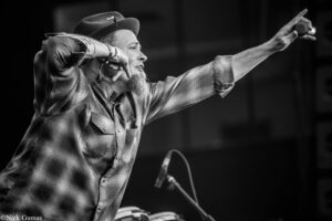 Bearded man in a plaid shirt and hat performing on stage, extending his right arm with a microphone toward the audience.