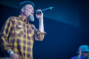 Male singer wearing a yellow plaid shirt and a blue hat, singing into a microphone on stage.